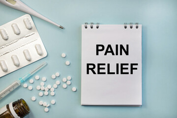 Pain relief is written on a notebook that lies among a thermometer and a blister with pills and a syringe. Medical concept.