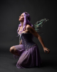 Full length portrait of a purple haired  girl wearing fantasy corset dress with fairy wings and flower crown.  Seated pose against a dark studio background.