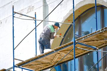 Construction worker on scaffolding with hammer clogs dowel into plastic umbrella, mount in insulation panel to the wall. Fixing rock wool insulation. Worker insulated facade standing at height