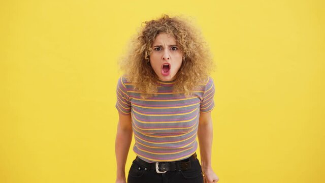 An angry curly blonde woman is screaming while standing isolated over yellow wall in the studio