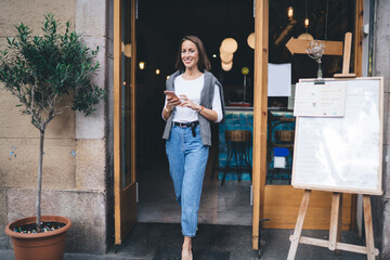Obraz na płótnie Canvas Full length portrait of cheerful female blogger with perfect smile holding modern cellphone device and looking at camera, cheerful girl with mobile device leaving coffee shop enjoying weekend time
