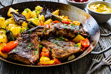 Tasty roasted ribs with baked potatoes and  vegetables served on frying pan on wooden table
