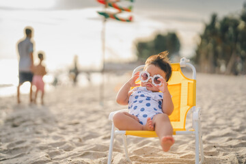 baby in white dress and sunglasses sit on the chair beach yellow color with relaxing and the wind...
