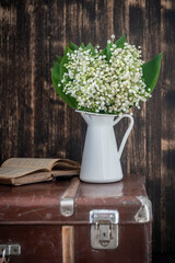 Beautiful bouquet of lilies of the valley in a white enamel vase on an old suitcase next to a book
