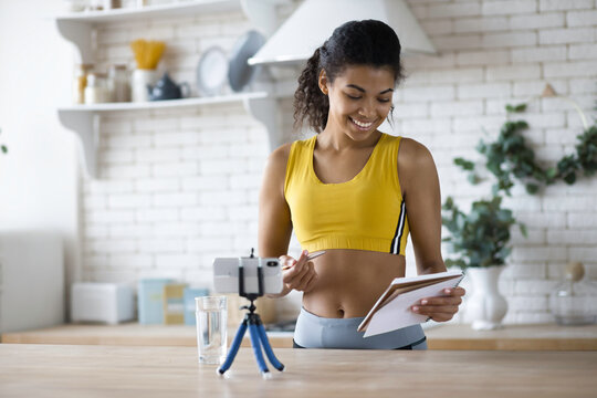 Young female blogger shoot social media vlog on smartphone, handsome African American woman fitness vlogger record streaming video about fitness and a healthy lifestyle at home in the kitchen - Powered by Adobe