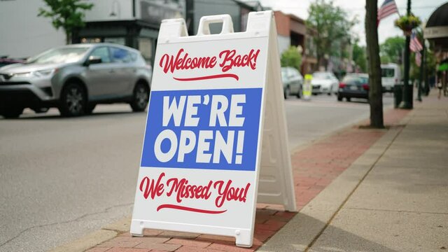 A sandwich board sign on the sidewalk outside of a small business on the main street of a small town proudly shows the store is now open. Businesses were hit hard during the Coronavirus pandemic of 20
