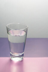 Vaso de cristal lleno de agua sobre una base de color rosa y lila. Vertical