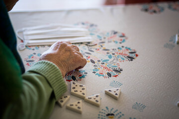 Main d'une femme âgée qui joue à un jeu de société sur une table napée, avec un masque dans le fond