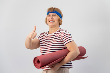 An elderly woman holds a yoga mat and shows her thumb up. The pensioner approves of sports.