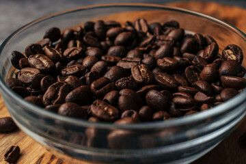 Fototapeta premium Coffee beans in a transparent bowl over a wood tablet with copy space. Dark background of coffee beans.