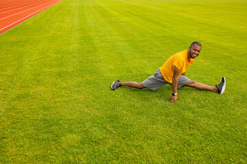 Horizontal shot of smiling active energetic African American male wearing sportswear and running shoes exercising at the stadium, doing splits on grass. Fitness, sports, activity and childhood