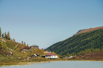 Fototapeta premium Weather station on hill near mountain lake in valley among high forest mountains. Scenic autumn landscape with mountain village on lake shore and mountains with coniferous forest. Three houses on hill