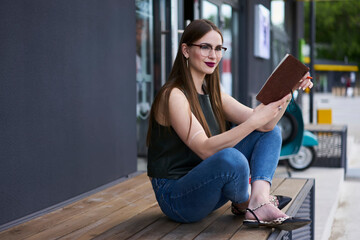 Pondering hipster girl in optical glasses for provide eyes correction thoughtful looking away while thinking about homework task, Caucasian female student with education textbook learning in city