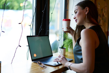 Millennial female copywriter with coffee to go reading web publication on modern laptop computer,...