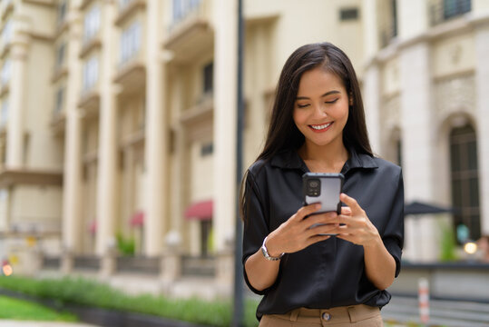 Asian businesswoman outdoors in city street using mobile phone while smiling and texting