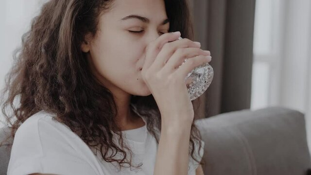 Cheerful young african american woman takes pill and drinks with glass of water in living room interior