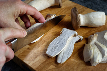 Close-up of King Oyster Mushrooms (Pleurotus eryngii) on a wooden cutting board. Man's hands guiding a chef's knife.