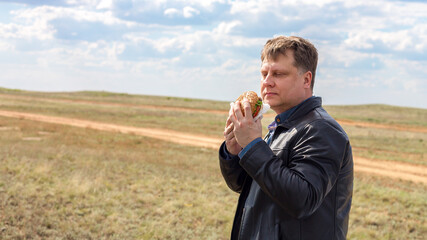 A hungry driver with a hamburger stands in the steppe, leaning on the car.