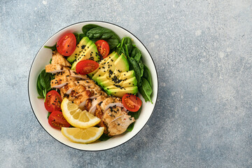 Fresh green salad with grilled chicken fillet, spinach, tomatoes, avocado, lemon and black sesame seeds with olive oil in white bowl on light slate background. Nutrition Diet Concept. Top view