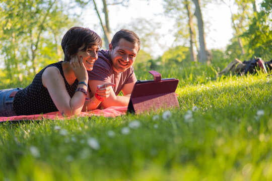 Photo of a young and attractive couple lying on a towel in the park watching a video on an electronic device and smiling at each other. Great summer day, relaxed attitude.