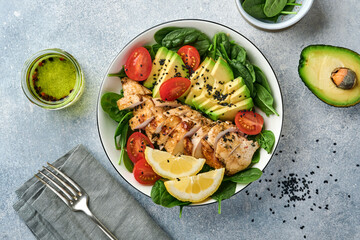 Fresh green salad with grilled chicken fillet, spinach, tomatoes, avocado, lemon and black sesame seeds with olive oil in white bowl on light slate background. Nutrition Diet Concept. Top view