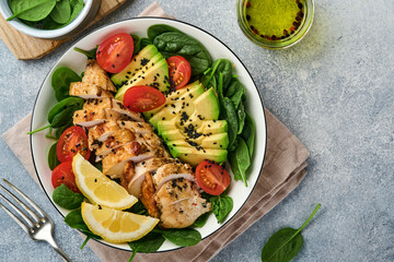 Fresh green salad with grilled chicken fillet, spinach, tomatoes, avocado, lemon and black sesame seeds with olive oil in white bowl on light slate background. Nutrition Diet Concept. Top view
