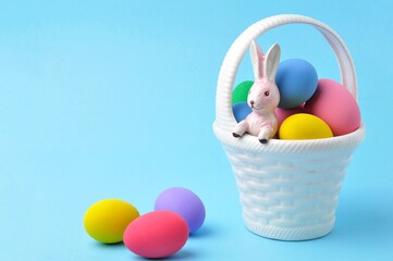 Several colored Easter eggs are on a white basket with a bunny inside on blue background.
