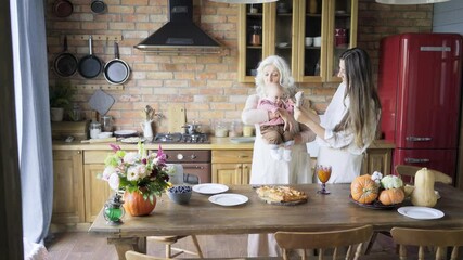 joyful aged lady hold little granddaughter in hands, kissing and hugging adult daughter near brown wooden kitchen table with pumpkin pie beverage and bouquet - Powered by Adobe
