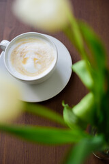 view through the flowers on the coffee with milk foam. white tulips and a cup and saucer on a wooden table. festive coffee break.