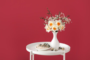 Vase with beautiful daffodils and quail eggs on table near color wall