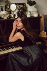 Young attractive lady is posing near piano, wearing black dress. Elegance