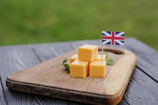 British cheddar cheese cubes on wooden board with United Kingdom flag. Man's hand places toothpick with flag of the UK - Powered by Adobe