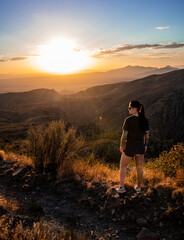 View of a young woman standing in the sunset looking out at mountains. She is a brunette with tattoo sleeves.
