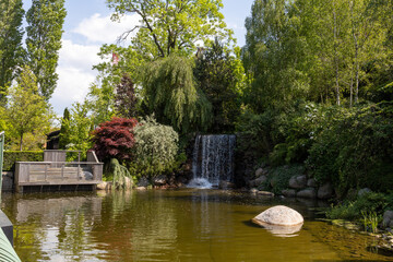 Pond with lush trees and bushes on sunny day. Small waterfall.