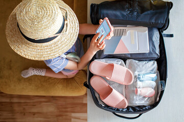 Pretty girl in straw hat and sunglasses sitting near suitcase packed for vacation and demonstrates...