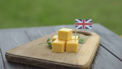 British cheddar cheese cubes on wooden board with United Kingdom flag. Man's hand places toothpick with flag of the UK