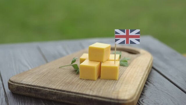 British cheddar cheese cubes on wooden board with United Kingdom flag. Man's hand places toothpick with flag of the UK