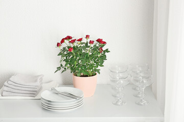 Obraz na płótnie Canvas Beautiful red roses in pot and tableware on shelf near light wall