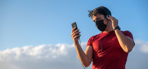 Naklejka premium Boy with mask, headphones and smartphone listening to music in the nature with blue skies