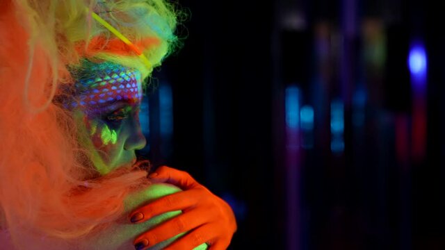 eccentric woman with fluorescent paint on face and body in nightclub, side portrait in darkness