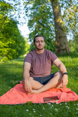 Naklejka na ściany i meble Photo of a young and attractive man sitting on a towel with his eyes closed and his legs crossed meditating in the middle of nature. Enjoying silence, relaxing with a guided meditation