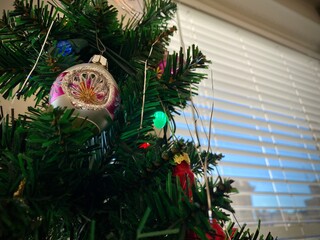 Ornaments, tinsel and lights on Christmas tree with blue sky visible through window blinds in...