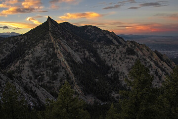 At the top of the Colorado flatirons during sunset. Mountain ridges with trees can be seen as well as a red, orange, yellow, and blue clouds.