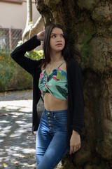 Fototapeta premium young female model with long hair posing standing by a tree in the city, wearing jeans and a colorful top, sunny summer day outside
