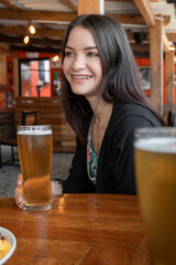 young woman with long straight hair sitting at a bar table with wooden decoration, holding a crystal glass with beer while smiling