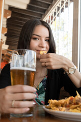 Young woman with long hair sitting in a restaurant, holding a drink and with the other eating a snacks, lifestyle and food