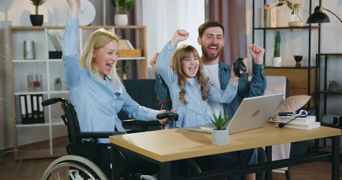 Family leisure concept where charming smiling happy light-haired woman in wheelchair after accident,her cute 10-aged daughter and loving bearded husband having fun together while playing videogames on