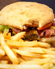 A partron has taken a bite out of a bacon cheeseburger garnished with tomato and lettuce.  French fries are blurred in the foreground.