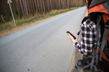 Female tourist in a check shirt with an orange large backpack near a highway in the woods with a smartphone in hand. Navigation, satellite maps, communication, domestic tourism. Backpacker, adventure 