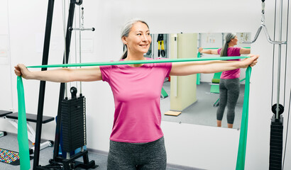 Fototapeta premium Patient woman doing exercises for recovery arms after injury using stretching resistance band at a rehab center